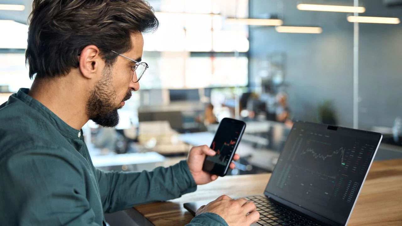 Man trading on his phone and laptop, analyzing forex charts to understand Bollinger Bands.